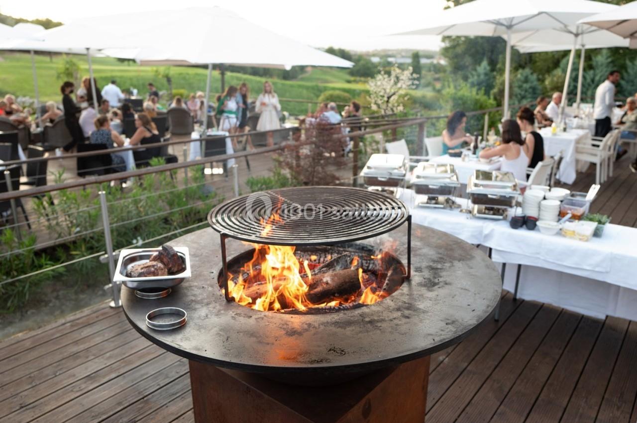 Barbecue circulaire en plein air avec flammes vives, entouré de tables dressées et de convives en arrière-plan.