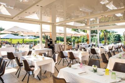 Salle de restaurant lumineuse avec tables rondes dressées, chaises marron et grandes baies vitrées.