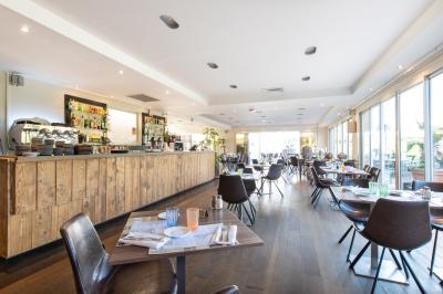 Salle de restaurant lumineuse avec tables rondes dressées, chaises marron et grandes baies vitrées.