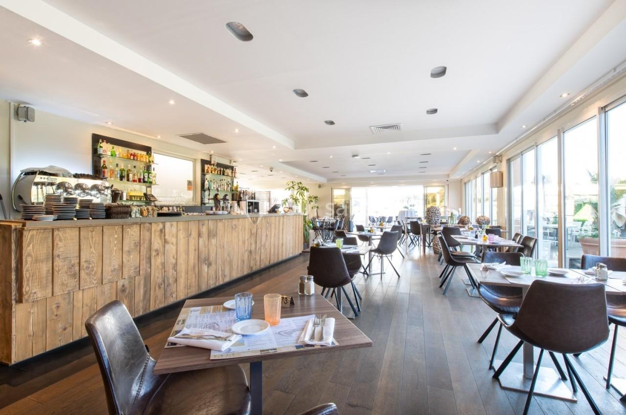 Salle de restaurant lumineuse avec un bar en bois, des tables dressées et de grandes baies vitrées.
