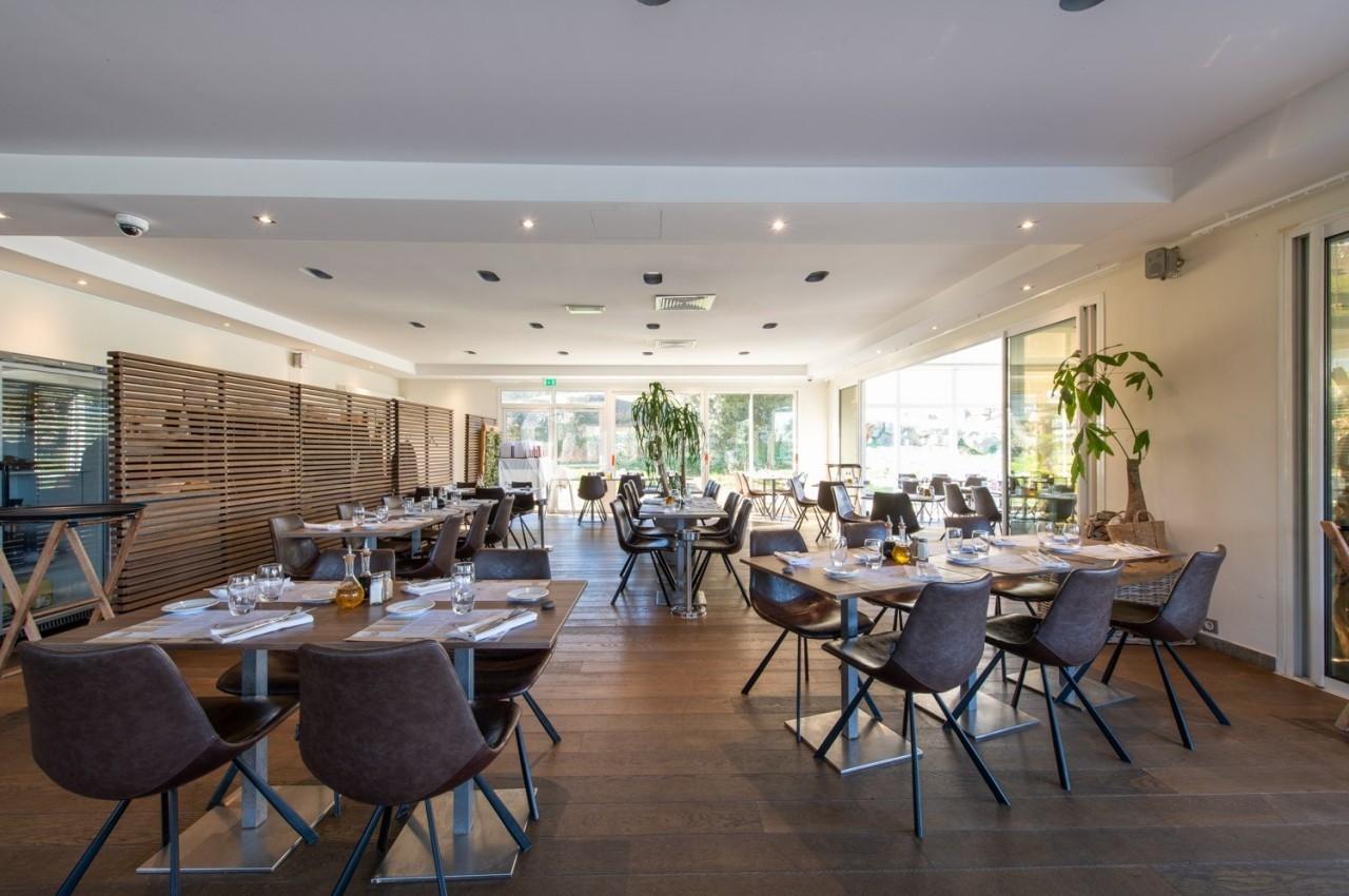 Salle de restaurant lumineuse avec tables dressées, chaises modernes et grandes baies vitrées donnant sur l'extérieur.