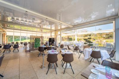 Salle de restaurant lumineuse avec tables rondes dressées, chaises marron et grandes baies vitrées.