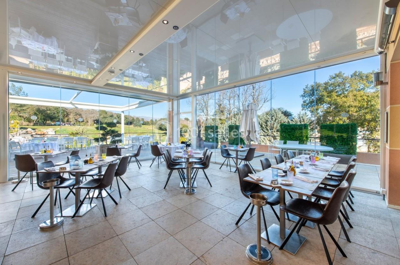 Salle de restaurant lumineuse avec grandes baies vitrées donnant sur une terrasse et un paysage verdoyant.