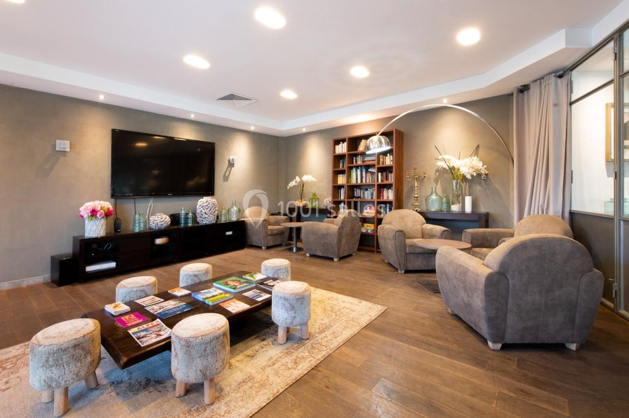Salon moderne avec fauteuils, table basse garnie de magazines, étagères de livres et écran mural, éclairage chaleureux.
