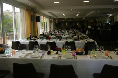 Salle de restaurant lumineuse avec tables rondes dressées, chaises marron et grandes baies vitrées.