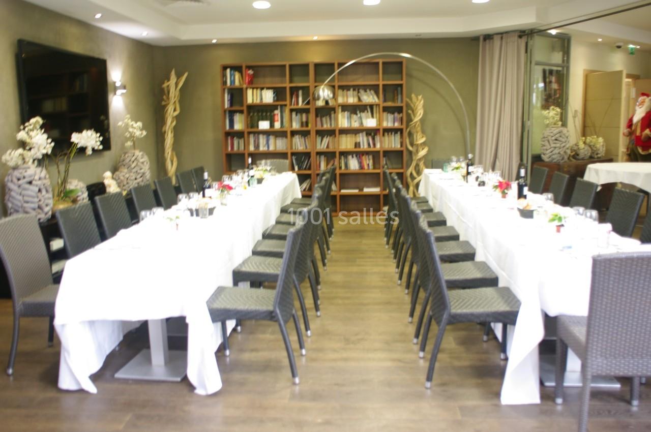 Salle de réception avec deux longues tables dressées, entourées de chaises noires, et une bibliothèque en arrière-plan.