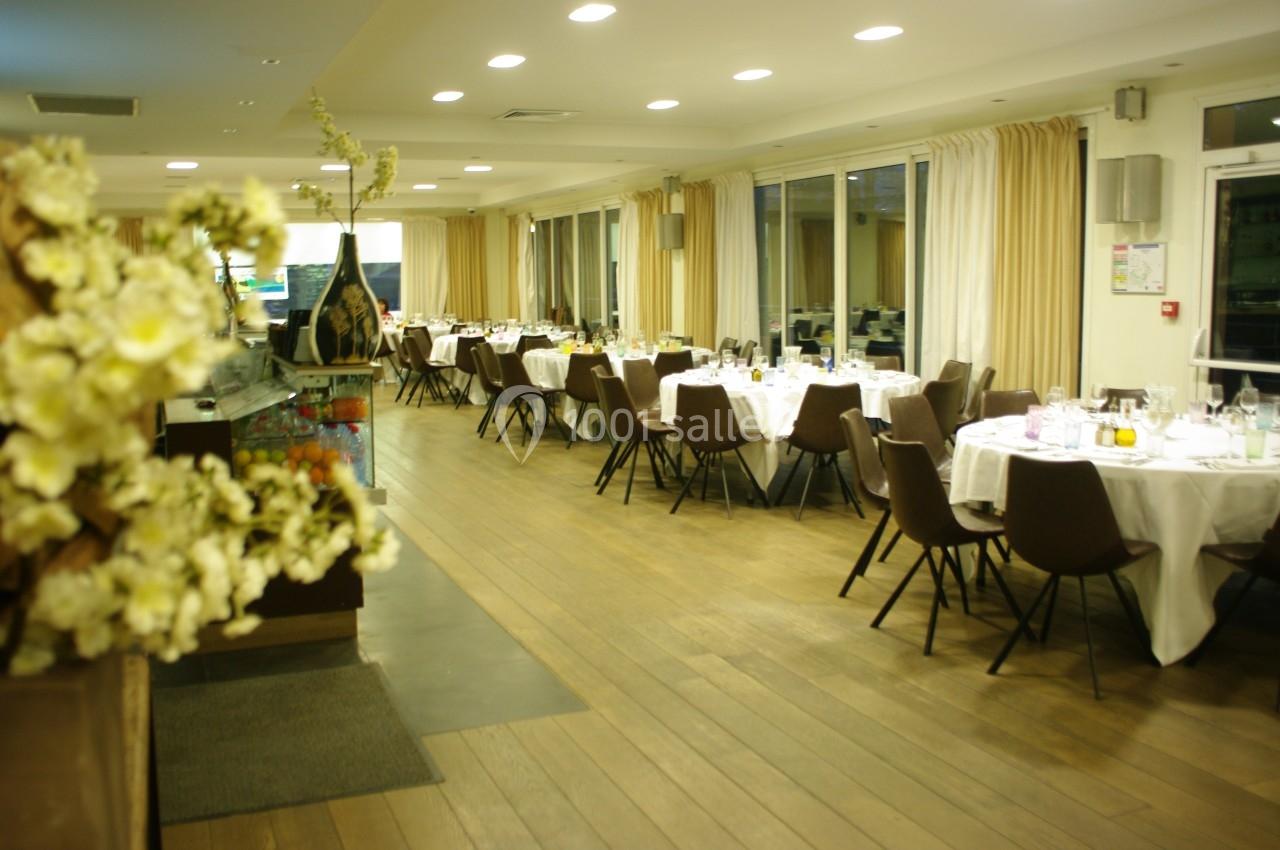 Salle de restaurant lumineuse avec tables rondes dressées, chaises marron et grandes baies vitrées.