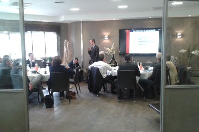 Salle de restaurant lumineuse avec tables rondes dressées, chaises marron et grandes baies vitrées.