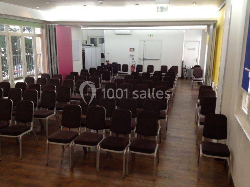 Salle de réunion avec des rangées de chaises vides alignées sur un sol en bois, éclairée par la lumière naturelle.