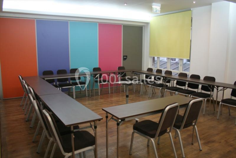 Salle de réunion avec tables disposées en U, chaises alignées et murs colorés en arrière-plan.