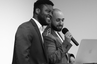 Deux hommes en costume parlent et sourient devant un ordinateur portable, l'un tenant un microphone.