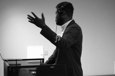 Un homme en costume, portant un casque audio, parle ou gesticule devant un pupitre dans une salle.