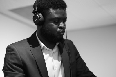 Un DJ en costume mixe de la musique devant une peinture représentant un village avec des collines en arrière-plan.
