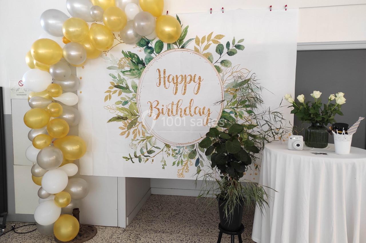 Décoration d'anniversaire avec arche de ballons dorés et blancs, panneau ’Happy Birthday’ et plantes en arrière-plan.
