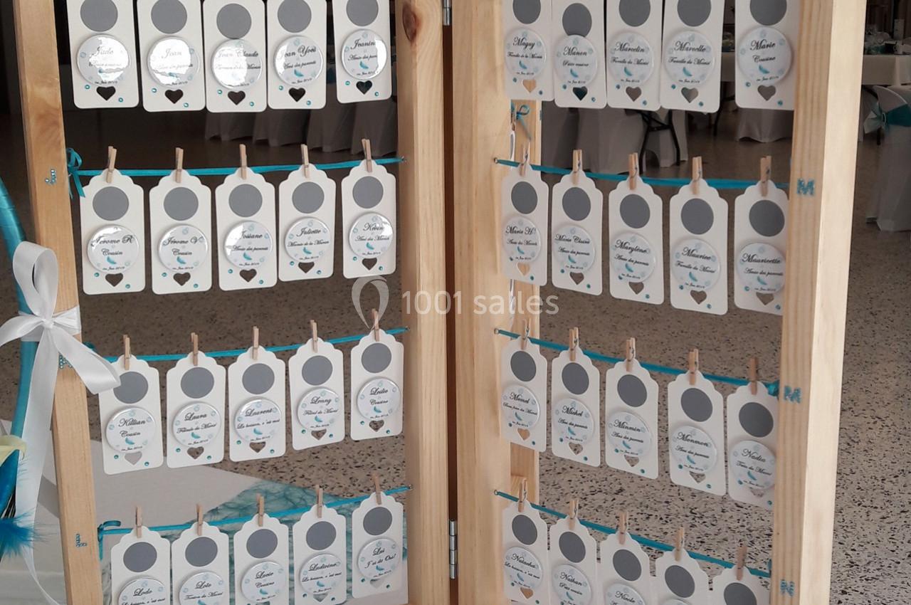 Panneau en bois avec des étiquettes blanches suspendues, affichant des noms et des tables pour un événement.