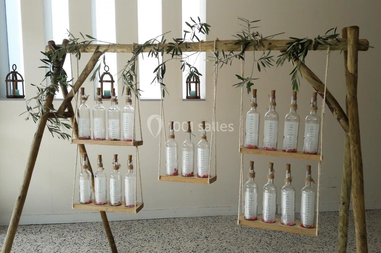 Bouteilles en verre transparent alignées sur des étagères suspendues à une structure en bois décorée de branches d'olivier.