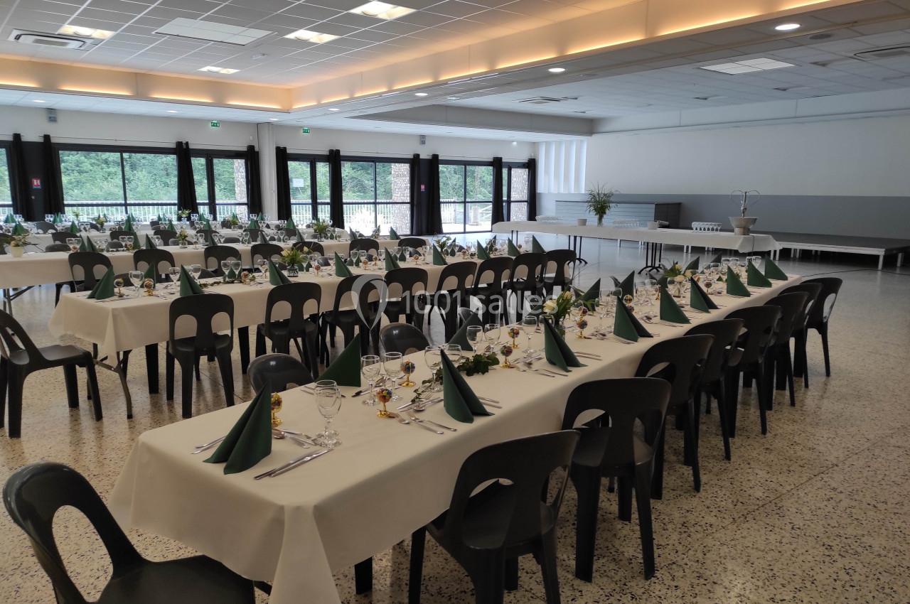 Salle de réception avec tables dressées, nappes blanches, serviettes vertes pliées et chaises noires.