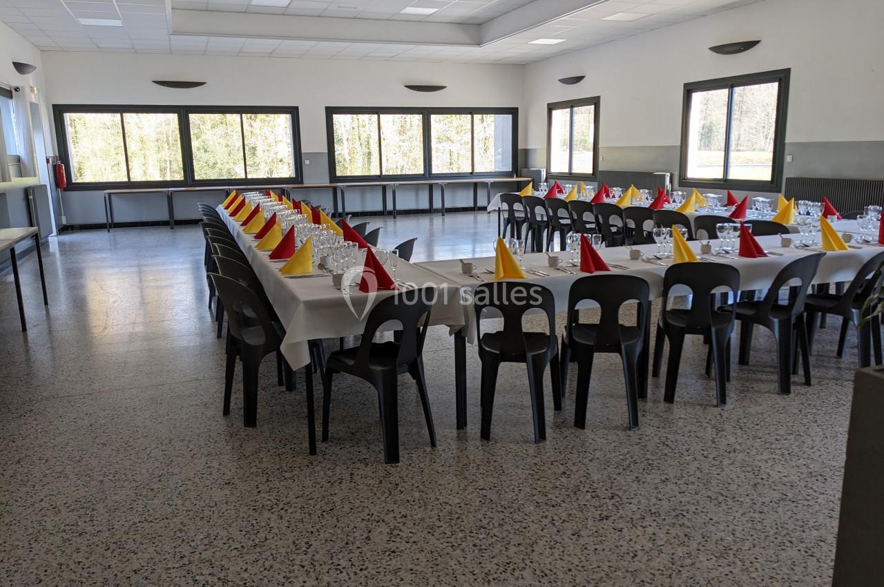 Salle de réception avec tables alignées, nappes blanches, chaises noires et serviettes pliées en rouge et jaune.