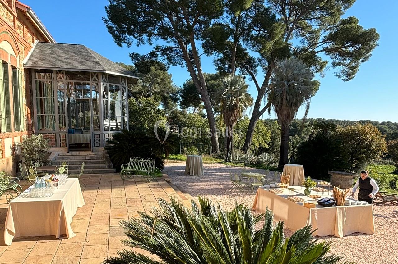 Terrasse ensoleillée avec tables dressées pour un buffet, entourée de végétation et d'arbres imposants.