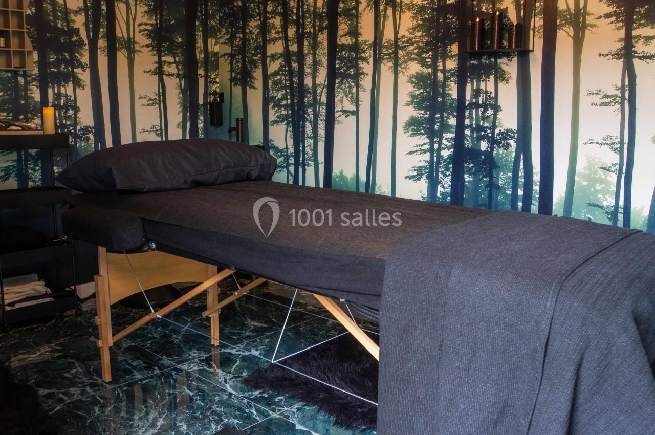 Table de massage recouverte de draps gris dans une pièce décorée d'un papier peint représentant une forêt.