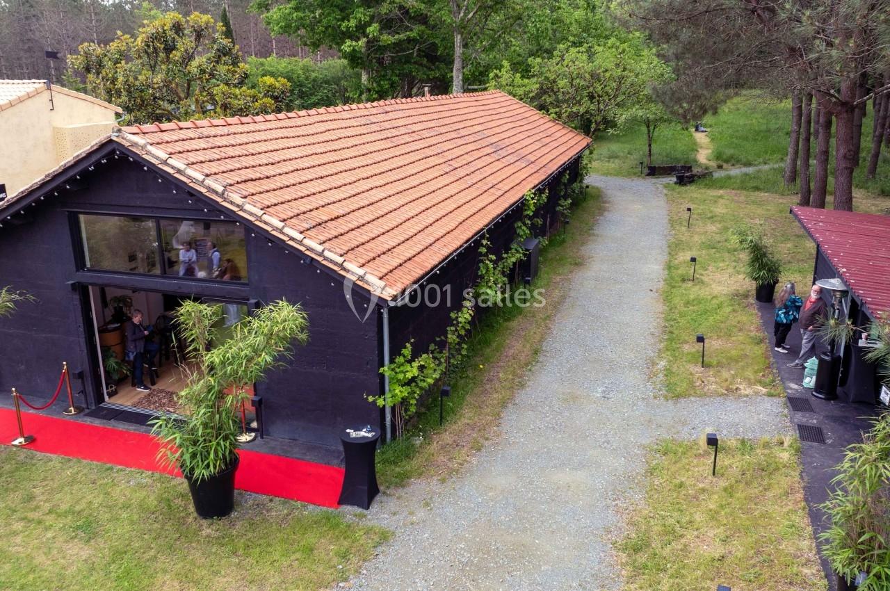 Bâtiment noir avec toit en tuiles rouges, entouré de végétation, avec une allée gravillonnée et une entrée vitrée.