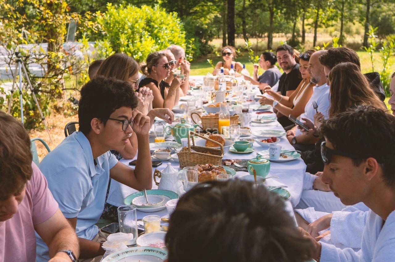 Groupe de personnes partageant un repas en plein air autour d'une longue table dans un cadre verdoyant.