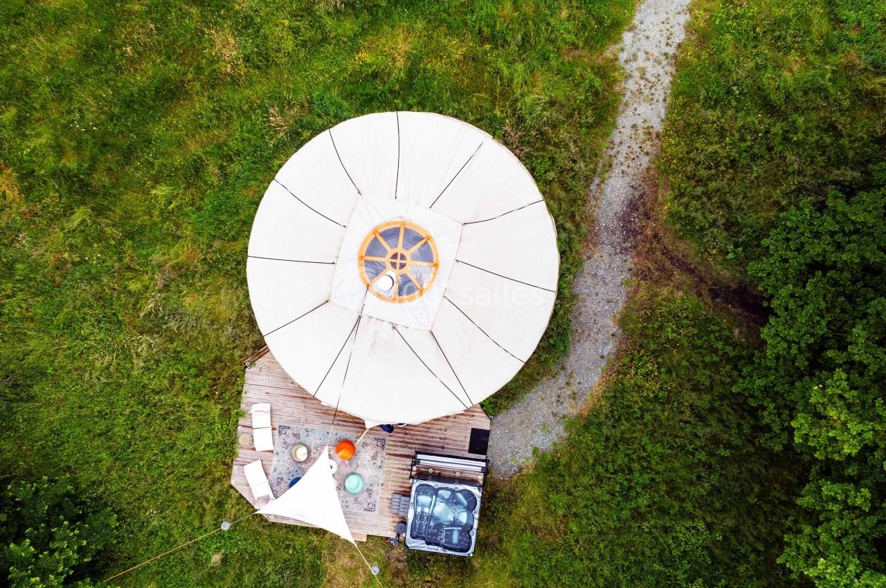 Vue aérienne d'une yourte blanche avec terrasse en bois, bain à remous et mobilier, entourée de verdure.