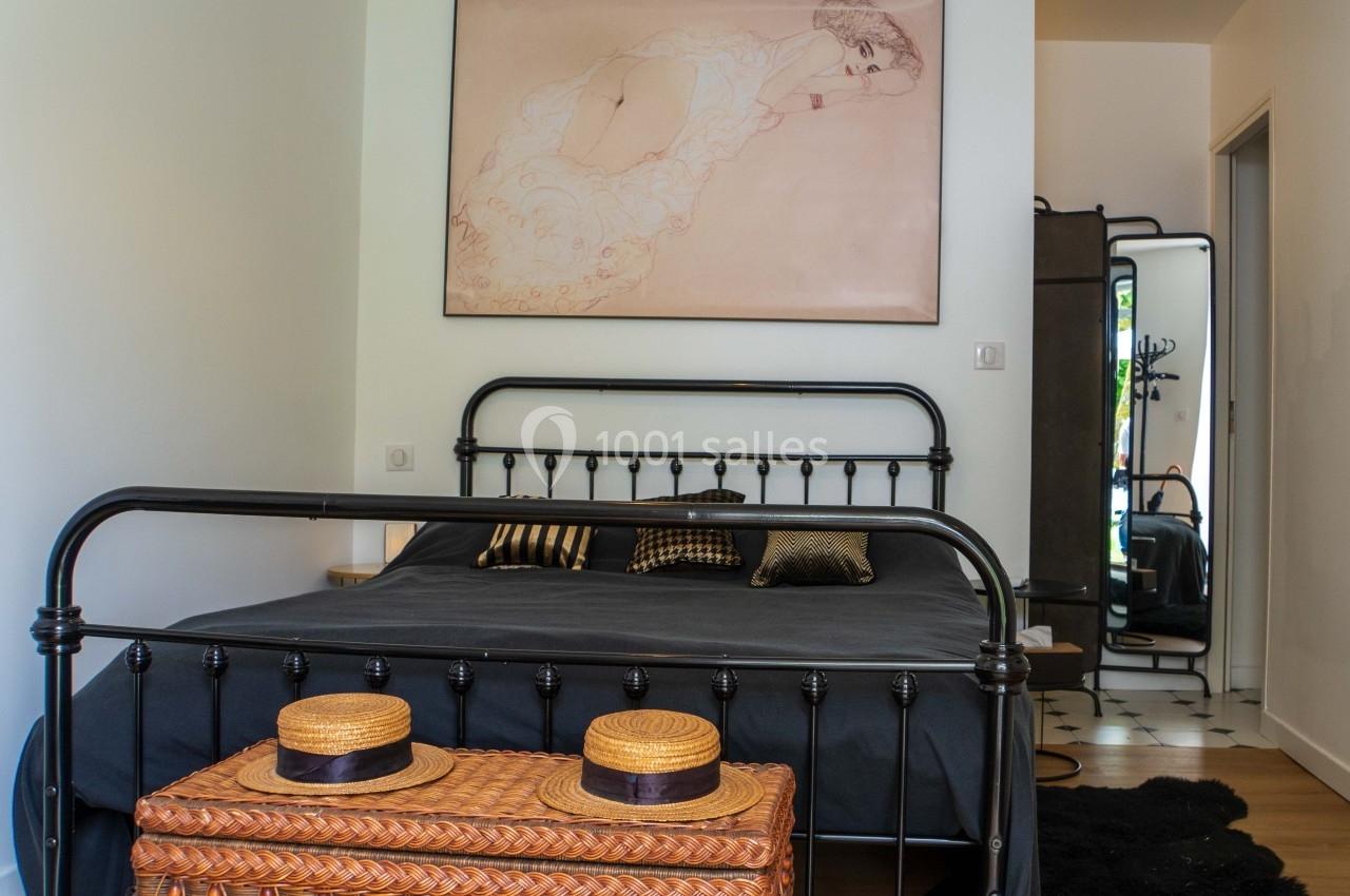 Chambre lumineuse avec lit en métal noir, coffre en osier au pied, et grand miroir dans un coin.