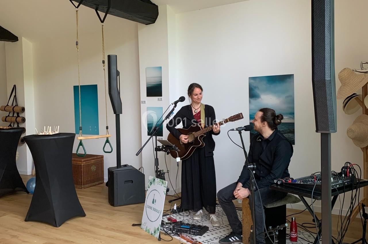 Deux musiciens jouent dans une pièce lumineuse avec des poutres apparentes, entourés d'équipements audio.