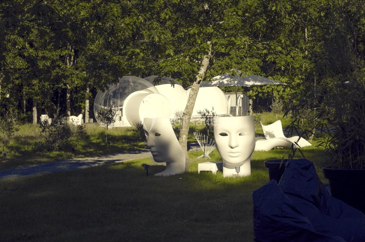 Sculptures de visages blancs dans un jardin verdoyant, avec des structures modernes en arrière-plan.