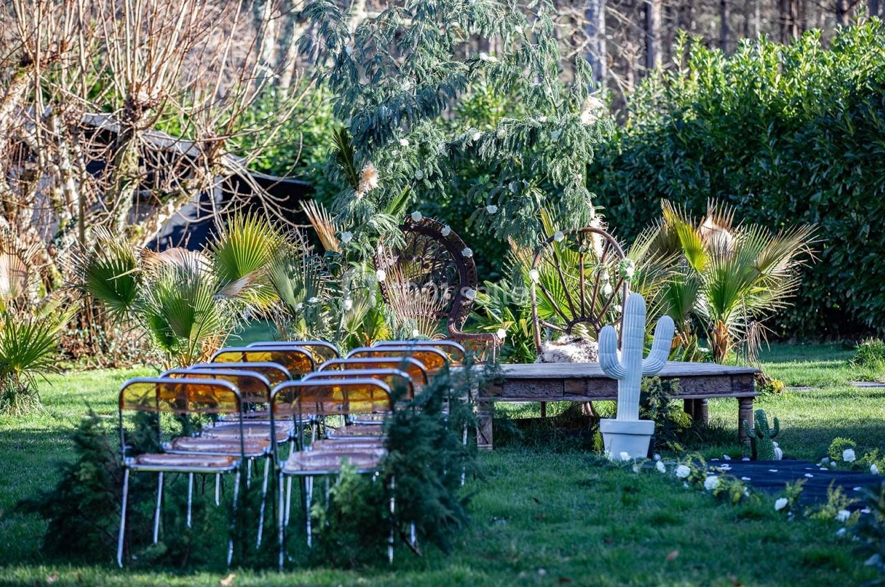 Chaises alignées sur une pelouse face à une table en bois décorée, entourée de plantes et d'arbustes dans un jardin.