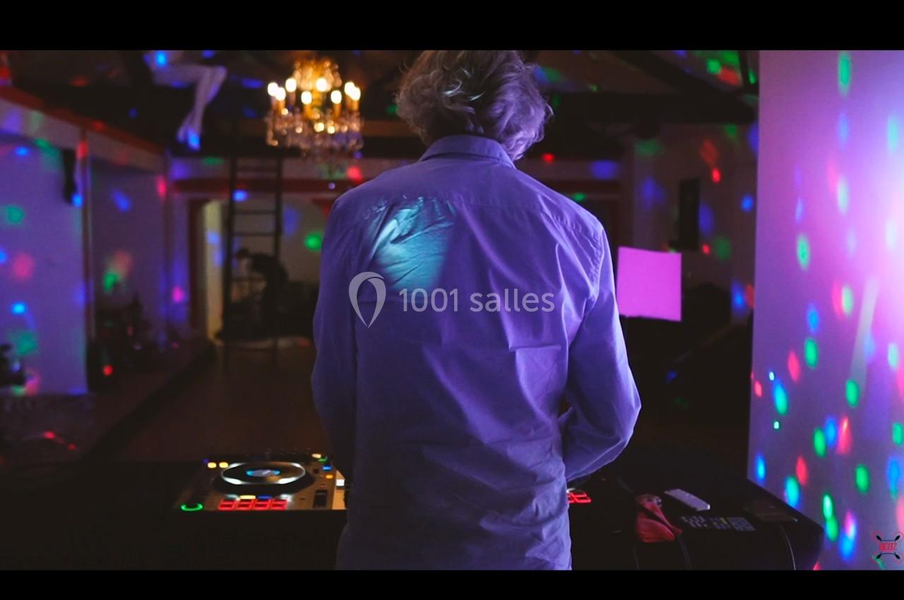 Un DJ vu de dos mixe dans une salle sombre éclairée par des lumières colorées et un lustre.