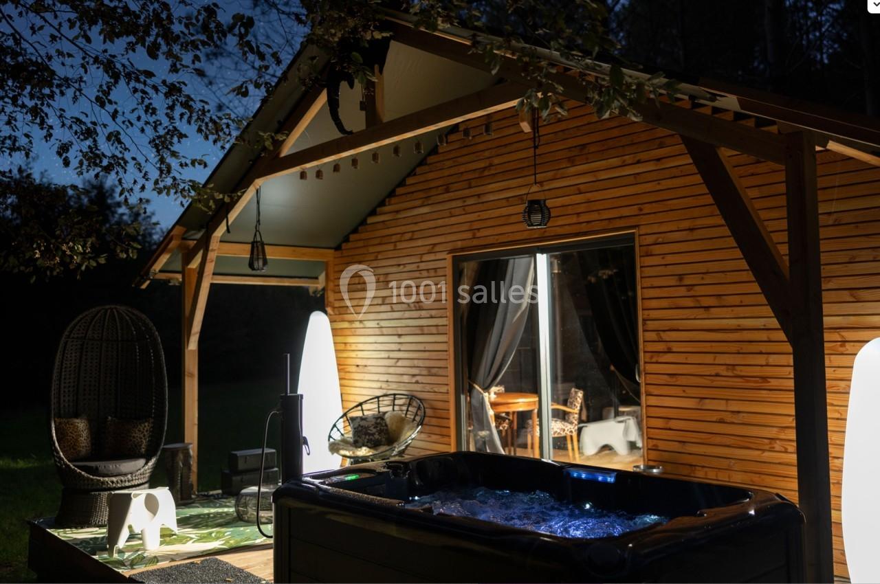 Chalet en bois éclairé de nuit avec terrasse aménagée, jacuzzi allumé et mobilier extérieur.