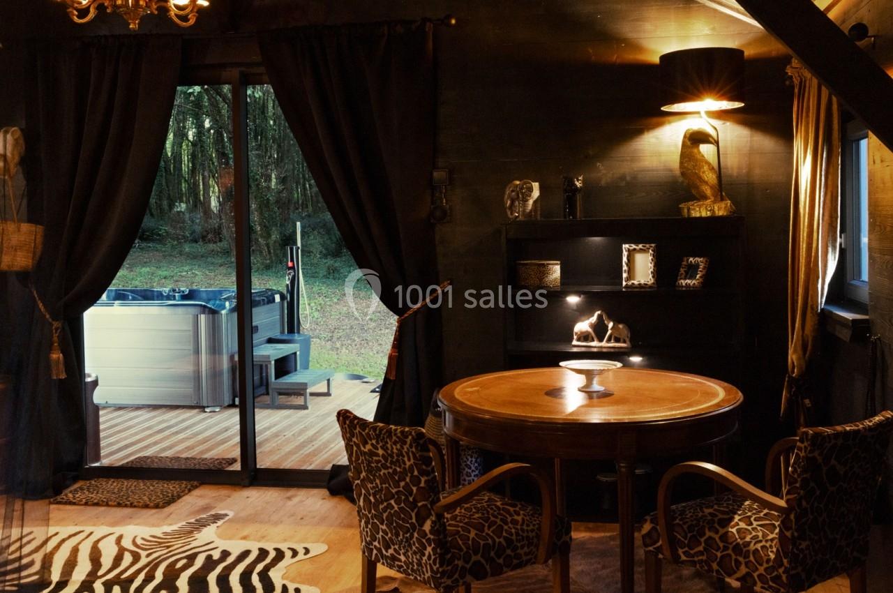 Intérieur chaleureux avec table ronde, fauteuils imprimés, et vue sur une terrasse avec un jacuzzi entouré de verdure.