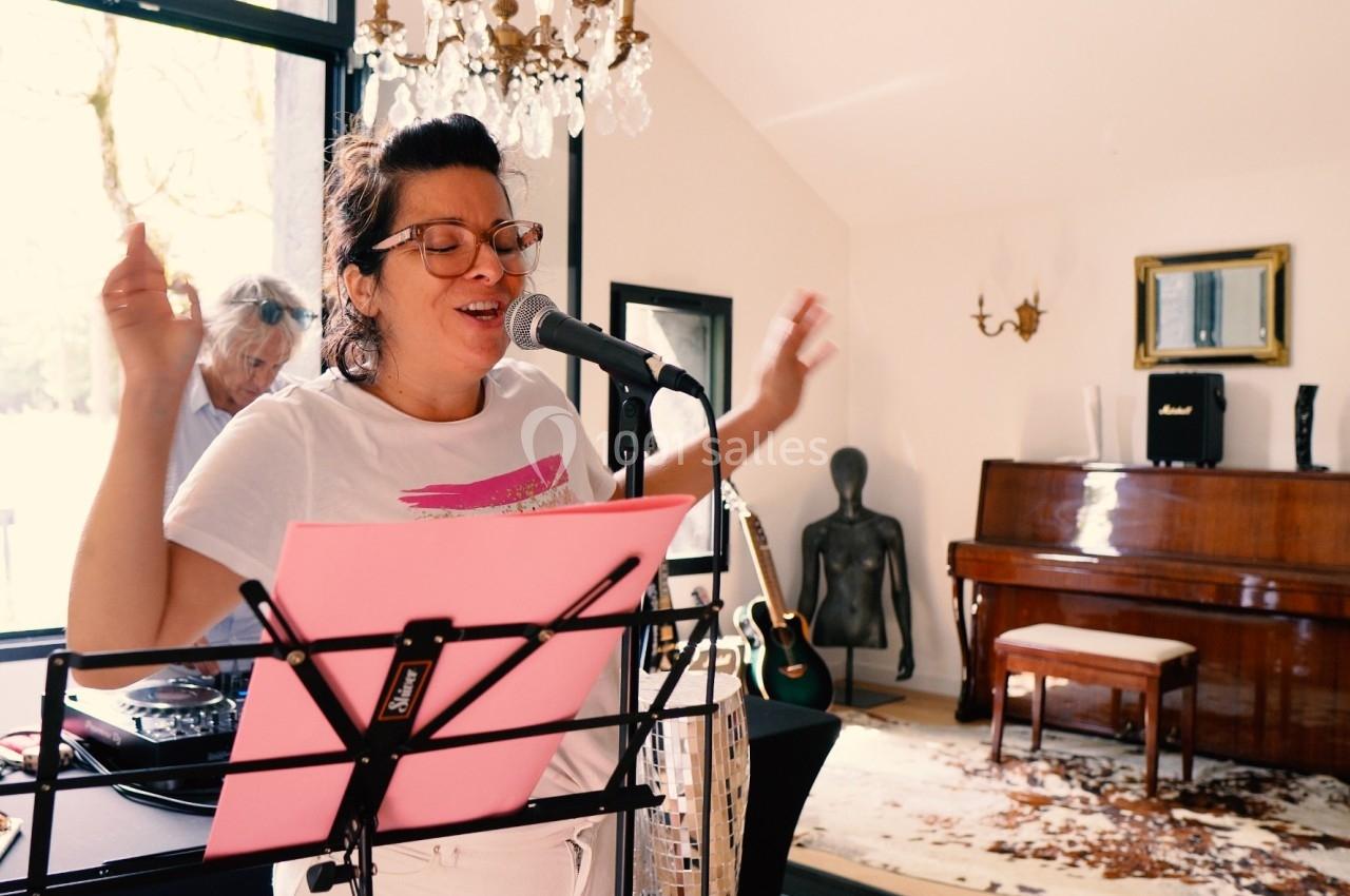 Une femme chante au micro dans une pièce lumineuse avec un piano, une guitare et un pupitre devant elle.