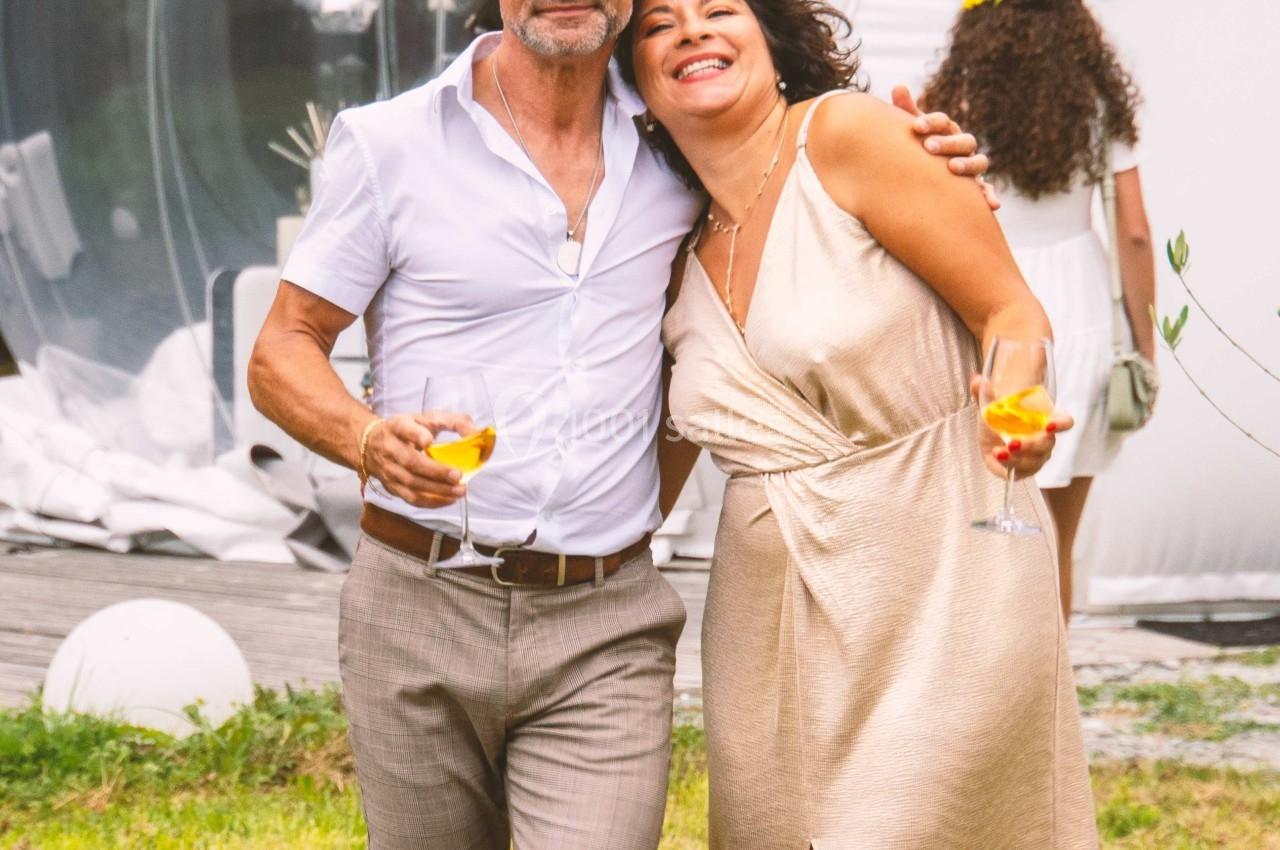 Un homme et une femme souriants tenant des verres, debout sur une pelouse devant une structure transparente.