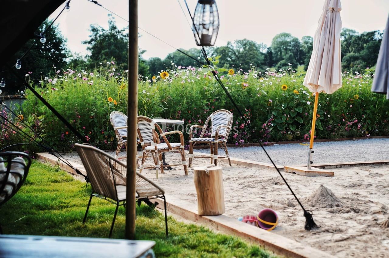 Chaises en rotin et table sur une terrasse en sable, entourées de fleurs et de verdure, avec un parasol fermé.
