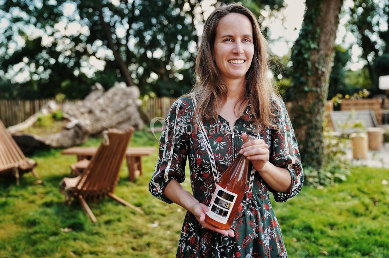 Une femme tenant une bouteille de boisson rosée dans un jardin avec des chaises en bois et des arbres en arrière-plan.