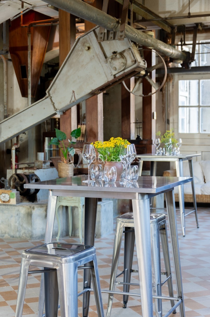 Tables hautes en métal avec verres et fleurs jaunes, dans un espace industriel lumineux au sol en carreaux.