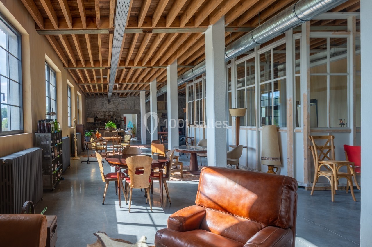 Espace lumineux avec plafond en bois, grandes fenêtres, mobilier varié et ambiance de loft industriel.