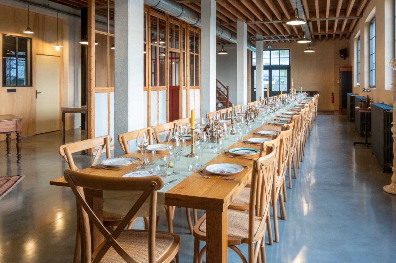 Grande table en bois dressée pour un repas, alignée avec des chaises dans une salle lumineuse au style industriel.