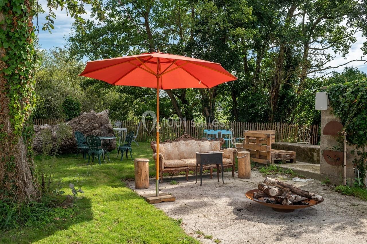 Espace extérieur avec canapé, parasol rouge, brasero et mobilier de jardin entouré de verdure.