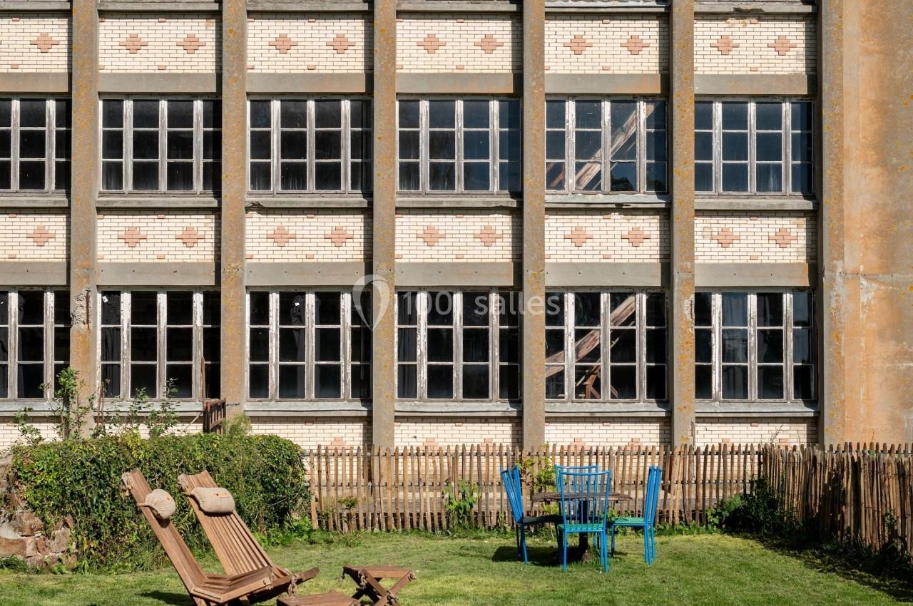 Façade d’un bâtiment en briques avec de grandes fenêtres, devant un jardin aménagé avec des chaises et des transats.