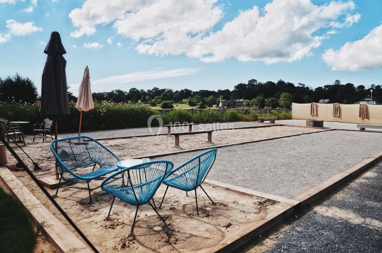 Terrain de pétanque en plein air avec deux fauteuils bleus, parasols et paysage champêtre en arrière-plan.