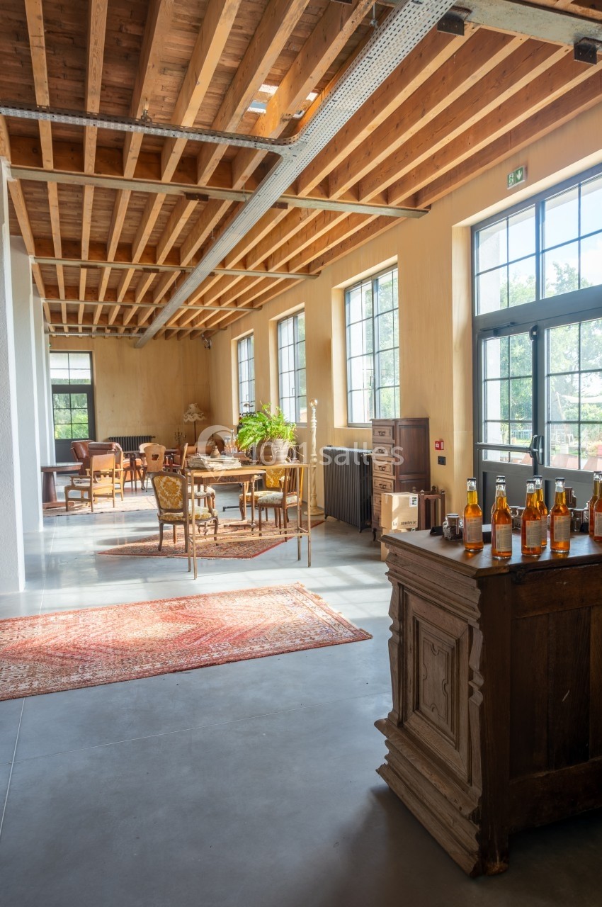 Salle lumineuse avec plafond en bois, grandes fenêtres, meubles anciens et bouteilles disposées sur un buffet.
