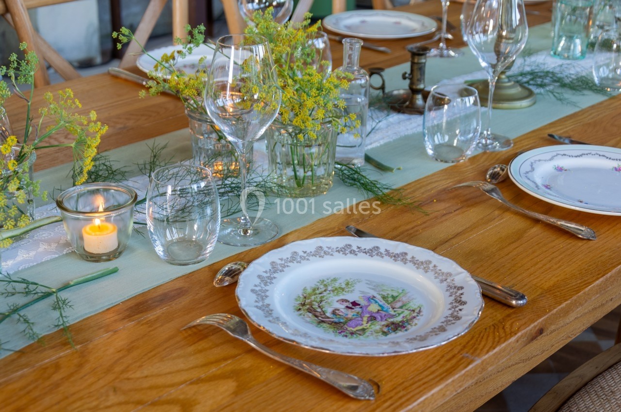 Table en bois dressée avec des assiettes décoratives, des verres, des couverts, des fleurs jaunes et une bougie allumée.