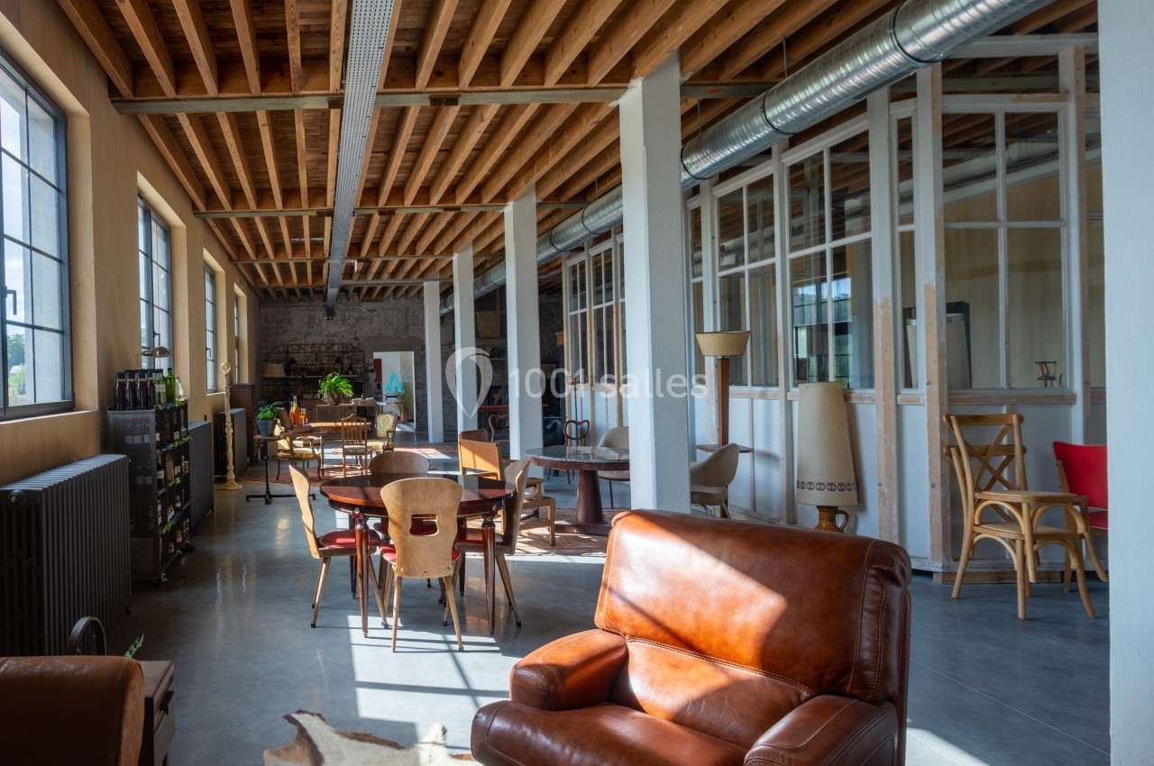 Espace lumineux avec mobilier en bois et cuir, grandes fenêtres, plafond en poutres apparentes et sol en béton.