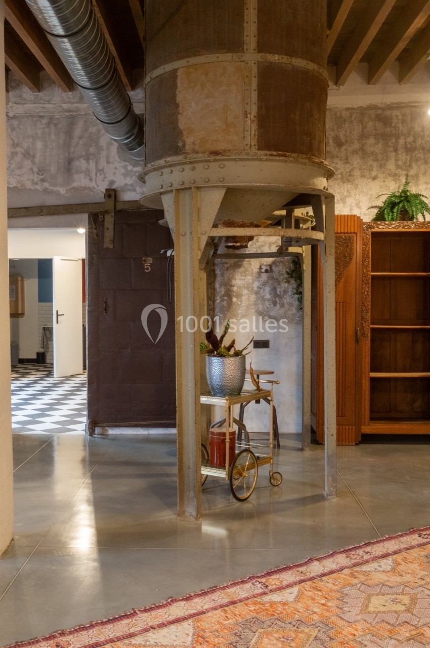 Ancien silo métallique intégré dans un intérieur industriel avec sol en béton, tapis coloré et mobilier en bois.