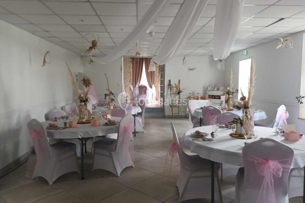Salle décorée pour un événement, avec tables rondes ornées de nappes blanches, chaises couvertes et touches de rose.