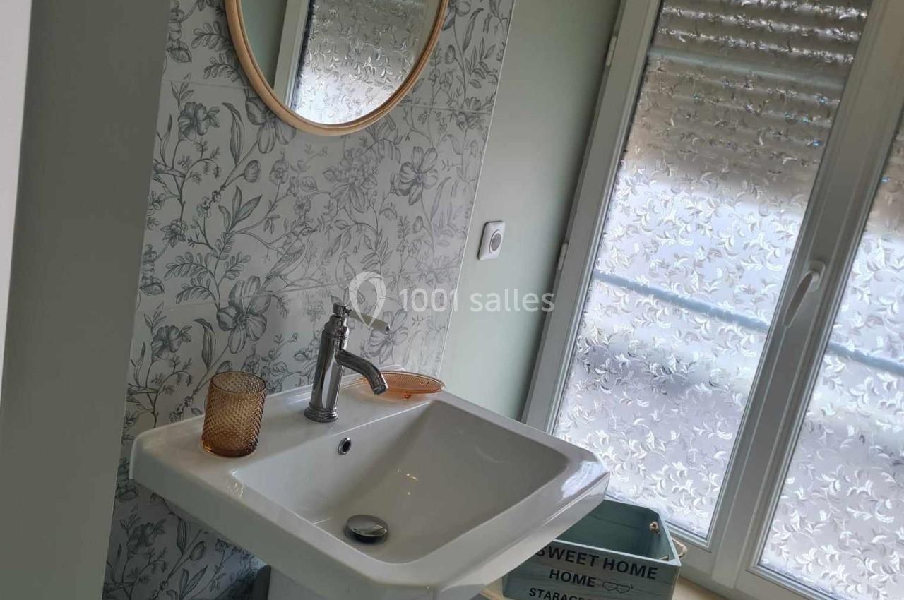 Lavabo blanc avec miroir rond, mur à motif floral, fenêtre avec stores baissés et étagère en dessous.